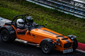 A bright orange open-top sports car with black racing stripes is speeding around a racetrack. Two individuals wearing helmets are seated inside, suggesting an experience focused on racing or thrill-seeking. The green grassy area and metal guardrail surround the track, indicating a controlled racing environment.