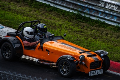 A bright orange open-top sports car with black racing stripes is speeding around a racetrack. Two individuals wearing helmets are seated inside, suggesting an experience focused on racing or thrill-seeking. The green grassy area and metal guardrail surround the track, indicating a controlled racing environment.