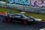 A sleek black sports car with red rims and a prominent rear spoiler is driving on a racetrack. The backdrop includes a fence lined with vibrant, colorful graffiti. The track surface shows some chalk or paint markings, suggesting a popular racing location.