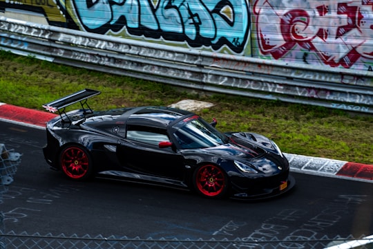 A sleek black sports car with red rims and a prominent rear spoiler is driving on a racetrack. The backdrop includes a fence lined with vibrant, colorful graffiti. The track surface shows some chalk or paint markings, suggesting a popular racing location.
