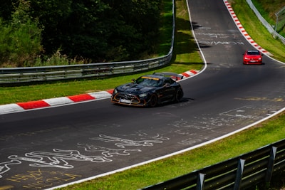 Two cars are driving on a racetrack, one black and one red, with greenery surrounding the track. The black car is ahead of the red car, and various graffiti and marks are visible on the asphalt. The track is winding with safety barriers on the sides.
