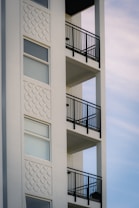 The image depicts a section of a modern building with an exterior wall featuring decorative white stucco patterns and a series of balconies with black metal railings. The structure exhibits a minimalistic design with clean, straight lines and large windows.
