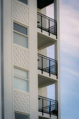 The image depicts a section of a modern building with an exterior wall featuring decorative white stucco patterns and a series of balconies with black metal railings. The structure exhibits a minimalistic design with clean, straight lines and large windows.