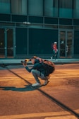 A person is crouched on a city street, holding a camera and appearing to take a photo, with a backpack on their back. The scene includes another person walking in the background near a building with large windows.