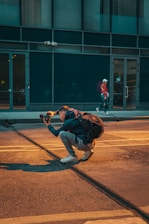 A person is crouched on a city street, holding a camera and appearing to take a photo, with a backpack on their back. The scene includes another person walking in the background near a building with large windows.