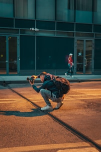 A person is crouched on a city street, holding a camera and appearing to take a photo, with a backpack on their back. The scene includes another person walking in the background near a building with large windows.