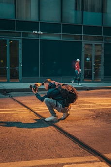 A person is crouched on a city street, holding a camera and appearing to take a photo, with a backpack on their back. The scene includes another person walking in the background near a building with large windows.