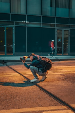 A person is crouched on a city street, holding a camera and appearing to take a photo, with a backpack on their back. The scene includes another person walking in the background near a building with large windows.