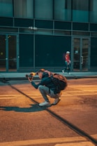 A person is crouched on a city street, holding a camera and appearing to take a photo, with a backpack on their back. The scene includes another person walking in the background near a building with large windows.