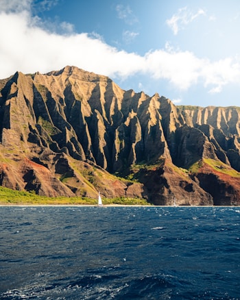 Towering, rugged cliffs rise dramatically from the ocean, showcasing a combination of earthy browns and lush greens. A small sailboat sails across the deep blue water, highlighting the vastness and grandeur of the natural landscape.