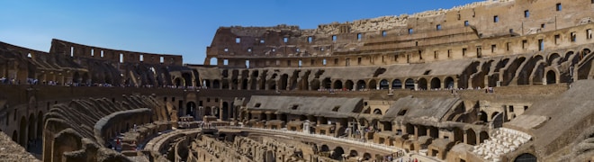 A panoramic view of the interior of an ancient amphitheater, showcasing the intricate stone arches, tiered seating, and weathered walls. The sunlit arena floor is partially visible with tourists scattered throughout the structure.