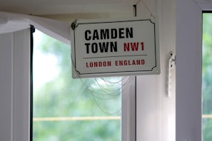 A vintage-style sign hangs indoors near a window, displaying the text 'CAMDEN TOWN NW1' in bold black letters with 'LONDON ENGLAND' in red beneath it. The sign is suspended by a string looped through two holes and positioned against a white-framed glass window. In the background, there is a soft focus view of green foliage, giving a glimpse of trees outside.