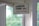 A vintage-style sign hangs indoors near a window, displaying the text 'CAMDEN TOWN NW1' in bold black letters with 'LONDON ENGLAND' in red beneath it. The sign is suspended by a string looped through two holes and positioned against a white-framed glass window. In the background, there is a soft focus view of green foliage, giving a glimpse of trees outside.