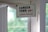 A vintage-style sign hangs indoors near a window, displaying the text 'CAMDEN TOWN NW1' in bold black letters with 'LONDON ENGLAND' in red beneath it. The sign is suspended by a string looped through two holes and positioned against a white-framed glass window. In the background, there is a soft focus view of green foliage, giving a glimpse of trees outside.