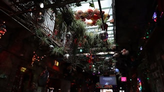 An indoor space with a glass ceiling decorated with hanging plants and disco balls. The atmosphere is vibrant and eclectic, featuring colorful lighting and a mix of natural and artificial elements, such as flowers and neon signs.