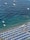 Aerial view of a beach filled with rows of blue and white umbrellas and sun loungers. The shoreline has people swimming in the clear, blue waters. Several boats and a floating platform are positioned further out in the sea.