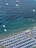 Aerial view of a beach filled with rows of blue and white umbrellas and sun loungers. The shoreline has people swimming in the clear, blue waters. Several boats and a floating platform are positioned further out in the sea.