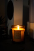 A lit scented candle glowing warmly in a dimly lit room. The candle is inside a translucent holder with a label on the front, and the surroundings have a cozy and intimate ambiance.