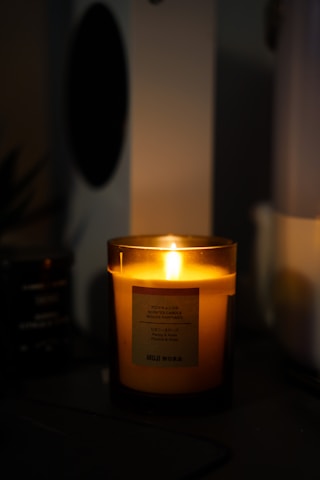 A lit scented candle glowing warmly in a dimly lit room. The candle is inside a translucent holder with a label on the front, and the surroundings have a cozy and intimate ambiance.