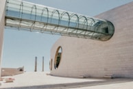 A modern architectural structure featuring a cylindrical glass walkway connecting two beige stone buildings. The building on the right has large circular windows and the sky is clear and blue. There are several benches placed sparsely in the open space.