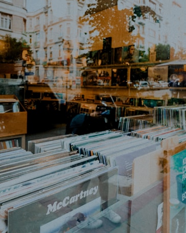 A record store window displays rows of vinyl records with covers of various artists. The reflection of a city street with buildings and trees is partially visible on the glass, blending the outdoor environment with the indoor shop setting.