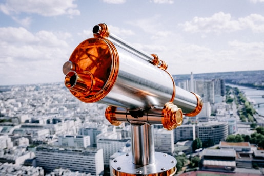 A metallic telescope with striking orange accents is mounted on a viewing platform, overlooking a vast, urban cityscape. The background features a river winding through the city, surrounded by numerous tall buildings under a partly cloudy sky.