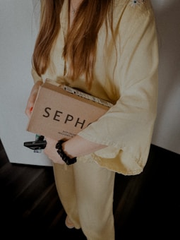 A person wearing a light yellow outfit holds a Sephora cardboard package in their arms. The individual has long brown hair and is wearing a black hair scrunchie on their wrist. The background appears to be an indoor setting with dark wooden flooring.