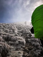 A group of hikers is ascending a frosted mountain path under a dark, overcast sky. The ground is covered with icy grass and rocks, creating a rugged terrain. The hikers are dressed in outdoor gear, with one wearing a bright green backpack, and they carry trekking poles.