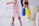 A person with a light blue shirt and beige pants holds two water bottles, one blue and one yellow, by their handle. In the background, a young girl wearing a pink dress and gray sneakers is visible.