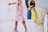 A person with a light blue shirt and beige pants holds two water bottles, one blue and one yellow, by their handle. In the background, a young girl wearing a pink dress and gray sneakers is visible.
