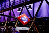 A vibrant, geometric structure illuminated with purple and blue lighting features a prominent Metro sign in the foreground. The sign has a red background with a blue label and white text. Spotlights shine downward, creating an urban, modern ambiance.
