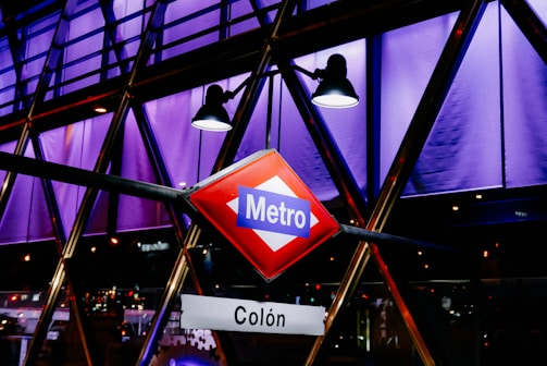 A vibrant, geometric structure illuminated with purple and blue lighting features a prominent Metro sign in the foreground. The sign has a red background with a blue label and white text. Spotlights shine downward, creating an urban, modern ambiance.