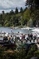 A crowd of people is gathered near a lake, with a background of dense green trees and several boats docked at a pier. The scene is lively, with people walking and engaging near parked cars, and a sign that says 'Queue Here.' The sky is partially cloudy, adding dimension to the natural setting.