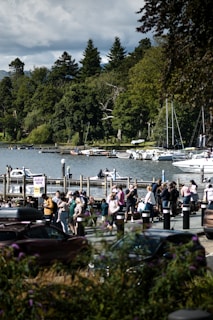 A crowd of people is gathered near a lake, with a background of dense green trees and several boats docked at a pier. The scene is lively, with people walking and engaging near parked cars, and a sign that says 'Queue Here.' The sky is partially cloudy, adding dimension to the natural setting.