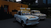 Two vintage cars adorned with colorful maps and signs are parked in front of a wall with a silhouette of a cityscape. The signs on the cars advertise sightseeing tours and indicate the next departure. The area is dimly lit, with an emphasis on the cars' decorative exteriors.
