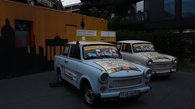 Two vintage cars adorned with colorful maps and signs are parked in front of a wall with a silhouette of a cityscape. The signs on the cars advertise sightseeing tours and indicate the next departure. The area is dimly lit, with an emphasis on the cars' decorative exteriors.