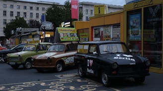 A row of vintage Trabant cars parked in front of a colorful information and souvenir building. The cars are painted in different colors and patterns, showcasing unique designs. The signage above displays flags of various countries, suggesting a tourist attraction. The surrounding environment includes urban buildings and greenery.