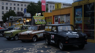 A row of vintage Trabant cars parked in front of a colorful information and souvenir building. The cars are painted in different colors and patterns, showcasing unique designs. The signage above displays flags of various countries, suggesting a tourist attraction. The surrounding environment includes urban buildings and greenery.