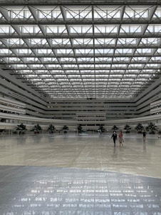 A spacious, modern indoor atrium with a high, grid-patterned ceiling allowing for ample natural light. The space features a large, polished floor and several large indoor plants adding greenery. People are scattered across the area, creating a sense of openness.