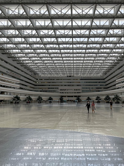 A spacious, modern indoor atrium with a high, grid-patterned ceiling allowing for ample natural light. The space features a large, polished floor and several large indoor plants adding greenery. People are scattered across the area, creating a sense of openness.