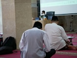 Three individuals seated on a red carpet in a room, facing a speaker at a table with microphones. The setting appears to be a formal or religious gathering, with the speaker wearing a face mask and a black cap.