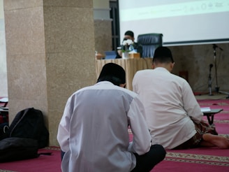 Three individuals seated on a red carpet in a room, facing a speaker at a table with microphones. The setting appears to be a formal or religious gathering, with the speaker wearing a face mask and a black cap.