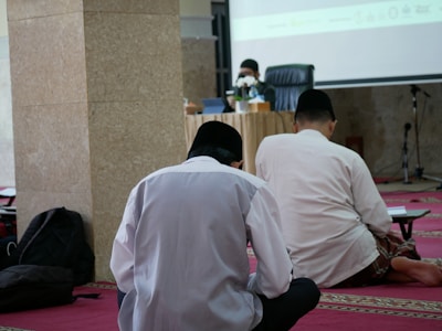 Three individuals seated on a red carpet in a room, facing a speaker at a table with microphones. The setting appears to be a formal or religious gathering, with the speaker wearing a face mask and a black cap.