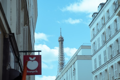 A narrow street lined with white residential buildings leads the eye to a distant view of the Eiffel Tower under a blue sky with scattered clouds. A red sign with a white apple icon and the word 'franprix' is visible in the foreground on the left.