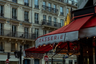 A red awning with the word 'BRASSERIE' is prominently displayed in front of a traditional European building. The building features ornate balconies and is constructed from light-colored stone. Warm lights hang under the awning, creating a cozy atmosphere. The scene captures the essence of a street corner in a European city.
