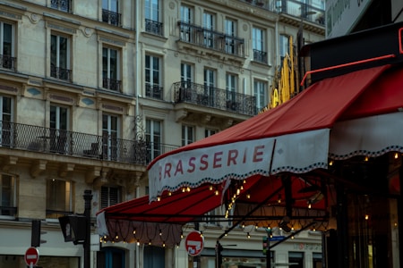 A red awning with the word 'BRASSERIE' is prominently displayed in front of a traditional European building. The building features ornate balconies and is constructed from light-colored stone. Warm lights hang under the awning, creating a cozy atmosphere. The scene captures the essence of a street corner in a European city.