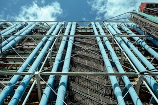 A large, architectural structure with numerous light blue pipes running vertically along a framework of scaffolding and metal supports. The perspective is upwards, revealing parts of the sky with scattered clouds in the background. The structure appears industrial, possibly part of a building's exterior.