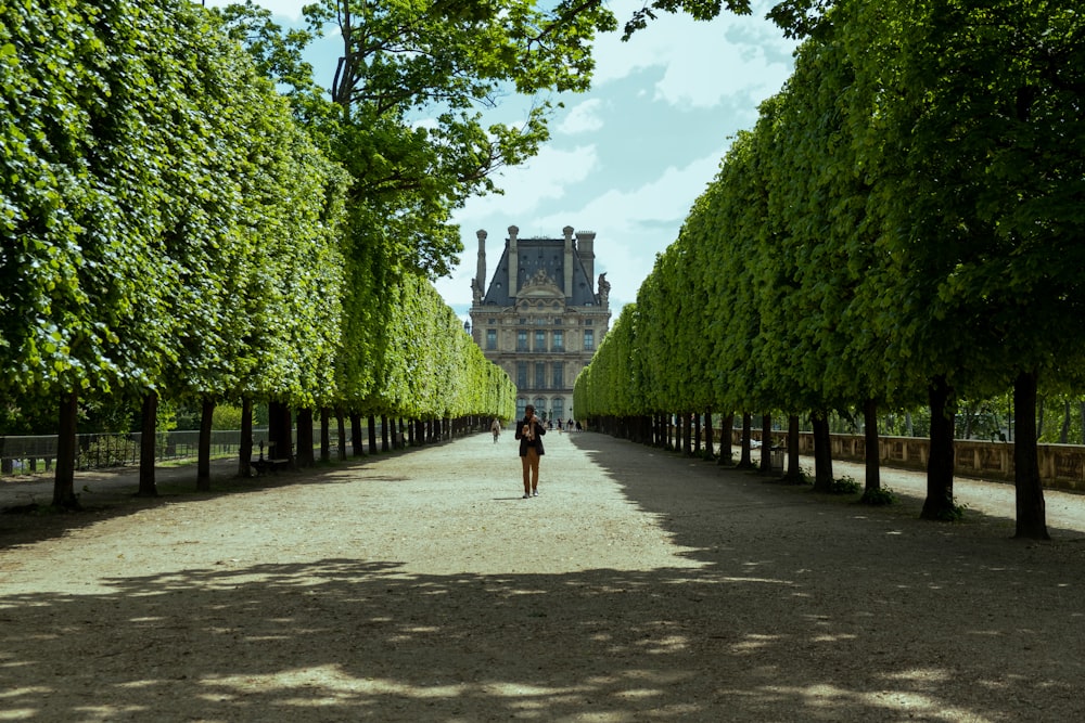 a woman walking down a dirt road next to tall trees