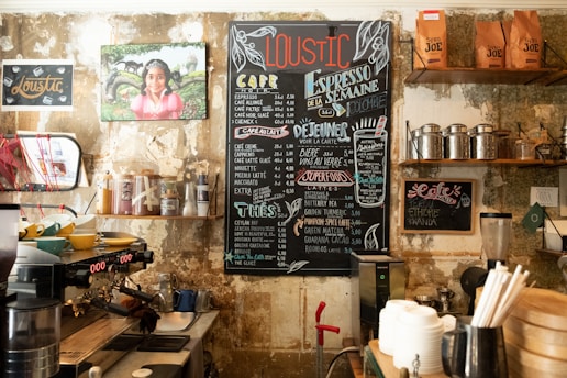 A cozy, rustic café interior with a prominently displayed chalkboard menu featuring various coffee and food options. The walls have a weathered texture adorned with creative artwork and photographs. Shelves host a variety of coffee-related items such as tins, bags of coffee beans, and cups. A professional espresso machine is situated on the countertop along with bowls and other café equipment.