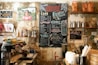 A cozy, rustic caf&eacute; interior with a prominently displayed chalkboard menu featuring various coffee and food options. The walls have a weathered texture adorned with creative artwork and photographs. Shelves host a variety of coffee-related items such as tins, bags of coffee beans, and cups. A professional espresso machine is situated on the countertop along with bowls and other caf&eacute; equipment.